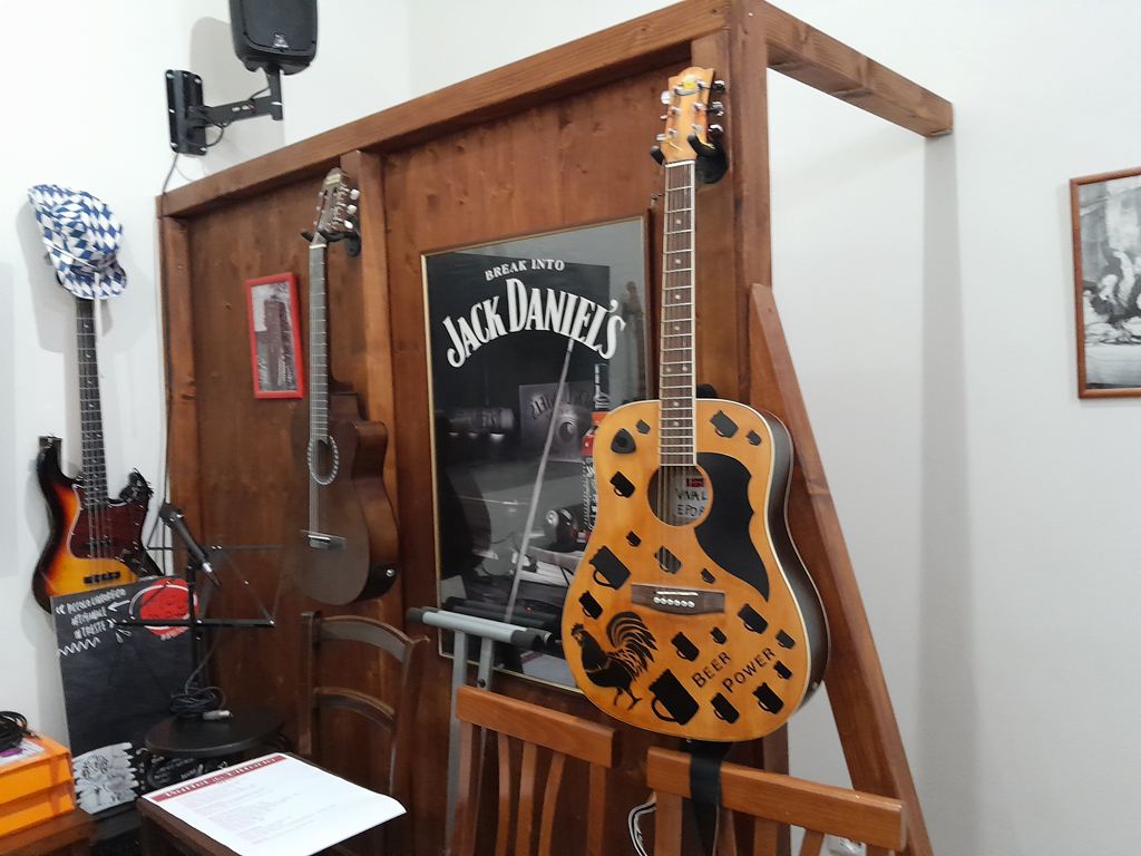 Una chitarra acustica decorata con un gallo, boccali di birra e la scritta «Beer Power», appesa a una parete di legno del Buffet da Vittorio accanto a una chitarra classica, un basso elettrico, una cornice rossa e una locandina di whisky