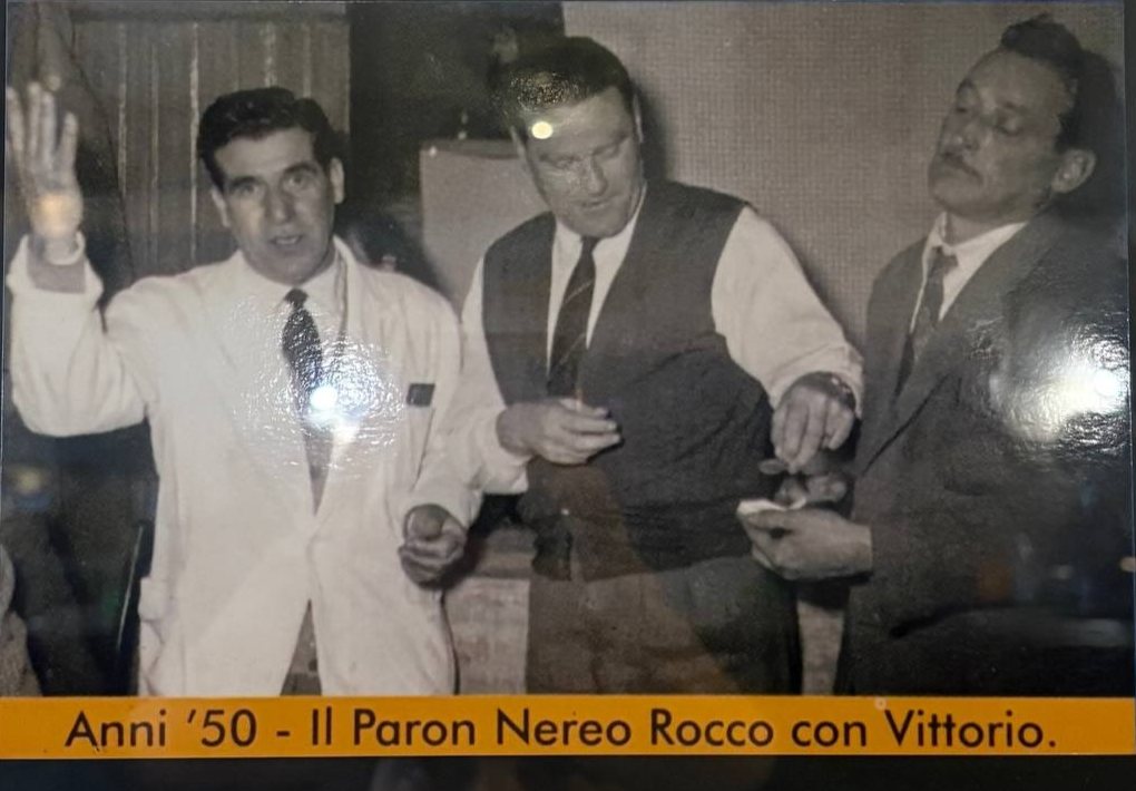 Fotografia in bianco e nero degli anni Cinquanta: tre uomini in piedi, uno in giacca bianca con la mano alzata, uno in gilet e cravatta e uno in giacca scura. La didascalia stampata dice «Anni '50 - Il Paron Nereo Rocco con Vittorio»